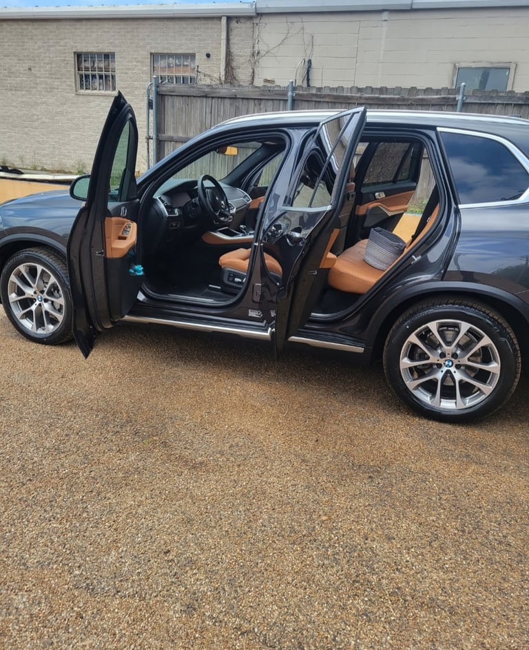Black SUV with tan/orange leather interior, open driver and rear doors, parked on gravel with industrial building background