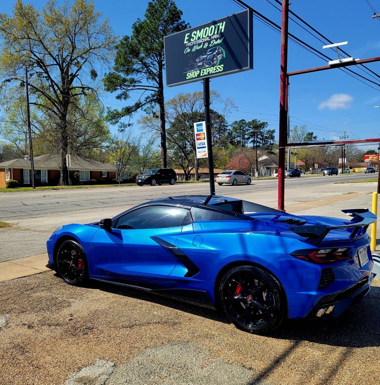 Bright blue sports car parked on a street under a shop sign with trees and commercial buildings in the background