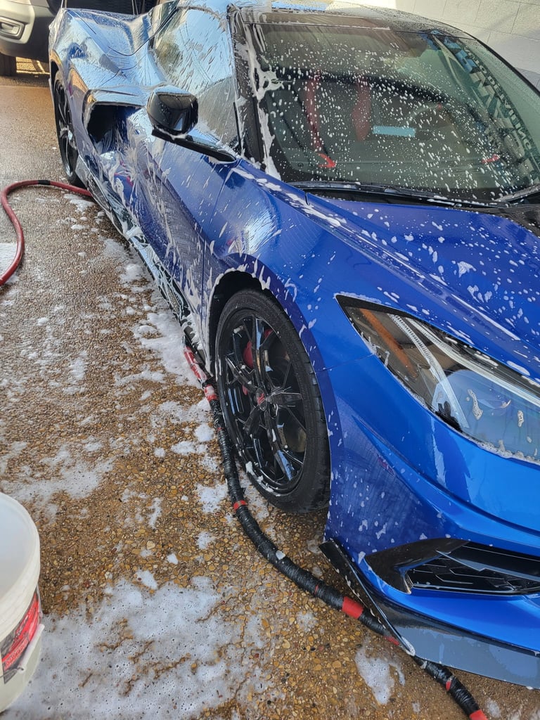 Blue car being washed with soap suds and water spray in an outdoor driveway with snow and ice on the ground