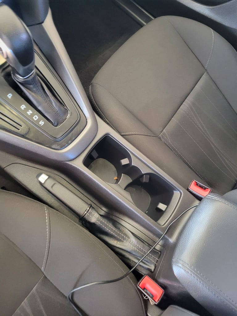 Car interior showing automatic transmission gear shifter, center console cup holder, and gray leather front seat with red seat belt buckles