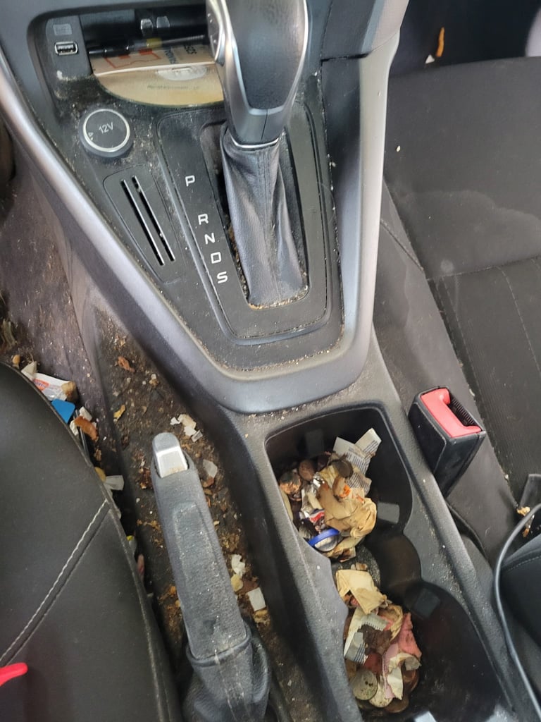 Car interior showing automatic transmission shifter and cup holders filled with trash and debris